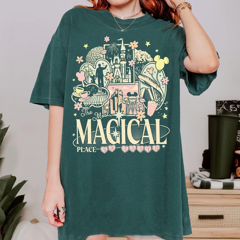 K&ouml;nnte beinhalten: Dunkelgr&uuml;nes T-Shirt mit einem farbenfrohen Aufdruck mit dem Text "The Most Magical Place on Earth". Die Grafik zeigt ein Schloss, Micky Maus und andere Themenparkelemente. Das Shirt hat kurze &Auml;rmel und einen Rundhalsausschnitt.