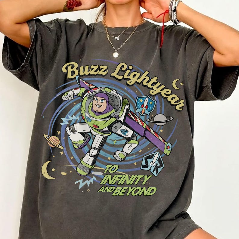 May include: A charcoal grey short-sleeve t-shirt featuring a graphic of Buzz Lightyear flying through space. The design includes the text Buzz Lightyear and To Infinity and Beyond, along with planets, stars, and a rocket ship emblem.