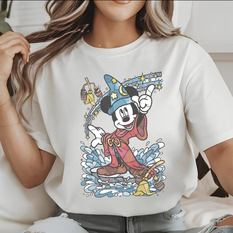 以下が含まれることがあります： ミッキーマウスが魔法使いに扮したカラフルなグラフィックが特徴のクリーム色のTシャツ。ミッキーは星の付いた青い魔法使いの帽子と赤いローブを着ています。デザインには星、水、ほうきが含まれています。