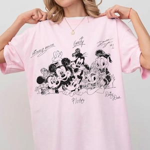 Puede incluir: Una camiseta rosa claro con un estampado gr&aacute;fico en negro estilo tinta de personajes de Disney, incluidos Mickey Mouse, Minnie Mouse, Goofy, Pluto, Donald Duck y Daisy Duck, con sus nombres escritos en cursiva encima o debajo de cada personaje.