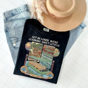 Puede incluir: Camiseta negra con el texto "GET IN LOSER WE'RE LEARNING ABOUT LETTUCE" y un gráfico de un paseo en barco. La camiseta se combina con vaqueros de lavado claro, un sombrero beige y un collar dorado.