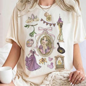 May include: Cream-colored t-shirt featuring a collection of cartoon illustrations. The design includes a princess portrait, a tower, a frying pan, a dress, and other related elements. The shirt has short sleeves and a round neckline.