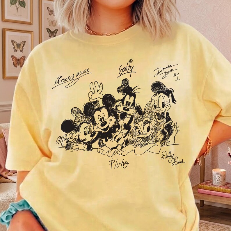 Puede incluir: Una camiseta de manga corta de color amarillo p&aacute;lido con un gr&aacute;fico de l&iacute;neas negras de personajes de Disney, incluidos Mickey Mouse, Minnie Mouse, Goofy, Pluto, Donald Duck y Daisy Duck, con sus nombres escritos en letra estilizada.