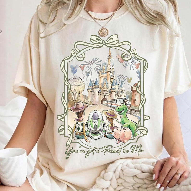 K&ouml;nnte beinhalten: Cremefarbenes T-Shirt mit einer gerahmten Illustration von Disney-Figuren vor einem Schloss. Die Illustration zeigt Woody, Buzz Lightyear, Rex und Hamm. Der Text "You've got a Friend in Me" steht darunter.