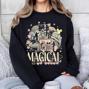 K&ouml;nnte beinhalten: Schwarzes Sweatshirt mit einem farbenfrohen Grafikdesign. Das Design zeigt ein Schloss, Micky Maus und die Worte "The Most Magical Place on Earth" in einer verspielten Schriftart. Rosa und gelbe Akzente erg&auml;nzen das Design.