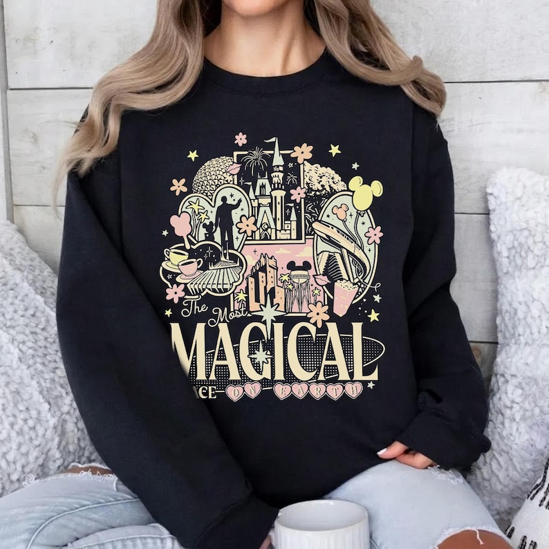 K&ouml;nnte beinhalten: Schwarzes Sweatshirt mit einem farbenfrohen Grafikdesign. Das Design zeigt ein Schloss, Micky Maus und die Worte "The Most Magical Place on Earth" in einer verspielten Schriftart. Rosa und gelbe Akzente erg&auml;nzen das Design.