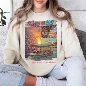 May include: Cream-colored sweatshirt with a colorful graphic featuring Mickey Mouse, Donald Duck, and other characters on a boat. The graphic includes the text "Keep it 100" and "on the land, the sea, the skies."
