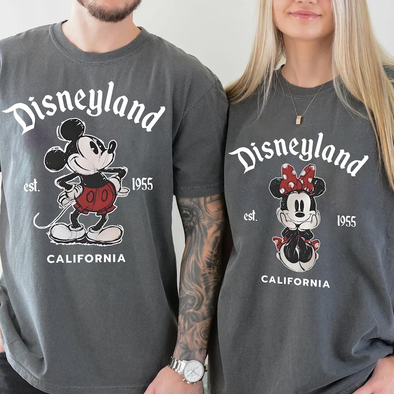 May include: Two gray t-shirts featuring the Disneyland logo with Mickey and Minnie Mouse graphics. The shirts have the text "Disneyland est. 1955 CALIFORNIA" in white. Mickey is wearing red shorts, and Minnie has a red bow.