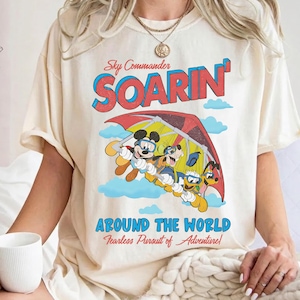 May include: A cream-colored t-shirt featuring a graphic of Mickey Mouse, Goofy, and Donald Duck hang gliding. The text reads Sky Commander SOARIN' AROUND THE WORLD Fearless Pursuit of Adventure in red and blue lettering against a sky background.