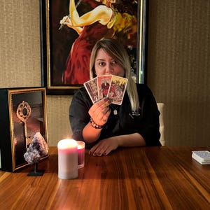 May include: A person holding three tarot cards, with a painting of a woman playing a violin in the background. A lit candle and a framed key are on the wooden table. A small stack of tarot cards is on the table.