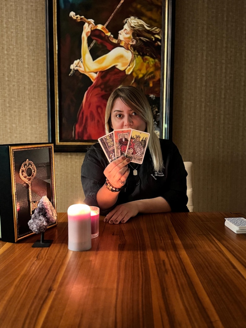 May include: A person holding three tarot cards, with a painting of a woman playing a violin in the background. A lit candle and a framed key are on the wooden table. A small stack of tarot cards is on the table.
