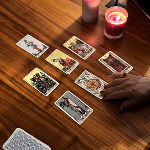 May include: A wooden table displays a tarot card reading. Several tarot cards are spread out, featuring illustrations and titles. A deck of cards is held in the foreground. Lit candles and decorative items are also present, creating an atmospheric setting.