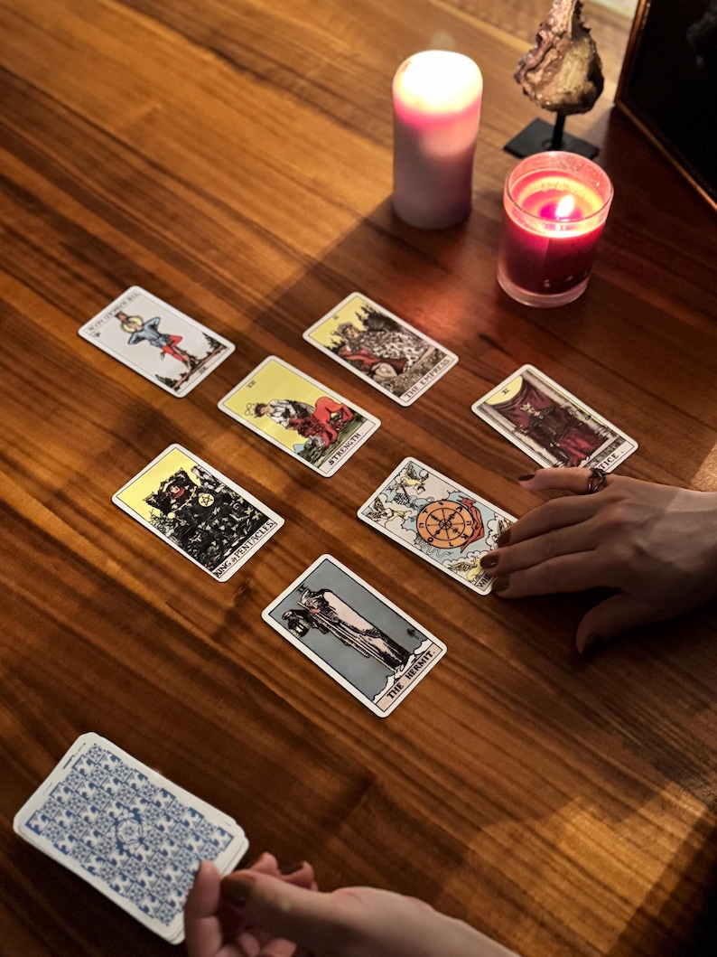 May include: A wooden table displays a tarot card reading. Several tarot cards are spread out, featuring illustrations and titles. A deck of cards is held in the foreground. Lit candles and decorative items are also present, creating an atmospheric setting.