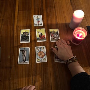 May include: A tarot card spread on a wooden table, featuring various cards with illustrations and titles. Two lit candles, one red and one white, are present. A hand with a bracelet is near the cards.