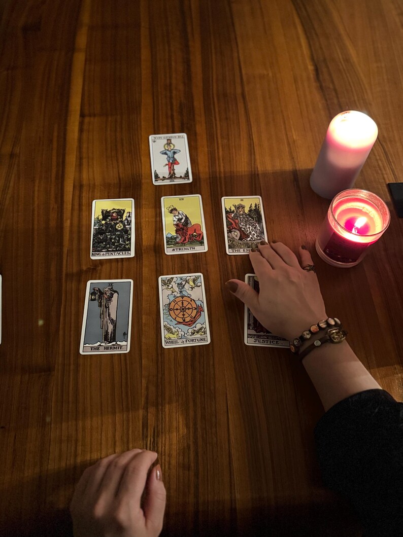 May include: A tarot card spread on a wooden table, featuring various cards with illustrations and titles. Two lit candles, one red and one white, are present. A hand with a bracelet is near the cards.