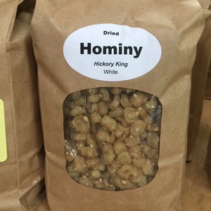 May include: A brown paper bag with a white label that reads "Dried Hominy Hickory King White". The bag contains dried hominy kernels.