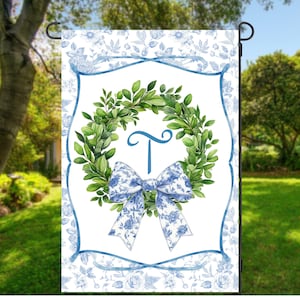 May include: A garden flag featuring a watercolor design. The flag has a white background with a blue floral pattern border. A green wreath with a blue bow and the letter "T" is in the center. The flag is displayed on a black metal stand.