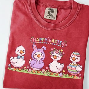 May include: A red Comfort Colors t-shirt featuring a "Happy Easter" graphic. The graphic includes four cartoon ducks, each with unique Easter-themed attire, standing on a green grassy area with Easter eggs and flowers.