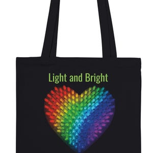 Puede incluir: Bolso tote negro con un diseño de corazón vibrante y de colores del arcoíris. El corazón está compuesto por numerosos puntos pequeños e iluminados. Las palabras "Light and Bright" están impresas en verde encima del corazón.