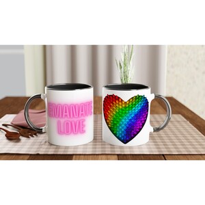 Puede incluir: Dos tazas de cerámica blanca con interior negro y asas plateadas. Una taza presenta el texto "EMANATE LOVE" en letras de neón rosa. La otra taza muestra un diseño de corazón de colores del arcoíris. Ambas tazas están sobre una mesa de madera.