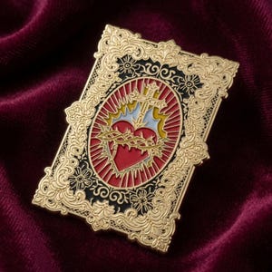 May include: A rectangular gold-toned metal pin featuring an ornate filigree border surrounding a central oval emblem. The emblem displays a red heart encircled by a crown of thorns, topped with a cross, set against a red and yellow sunburst.