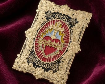 May include: A rectangular gold-toned metal pin featuring an ornate filigree border surrounding a central oval emblem. The emblem displays a red heart encircled by a crown of thorns, topped with a cross, set against a red and yellow sunburst.