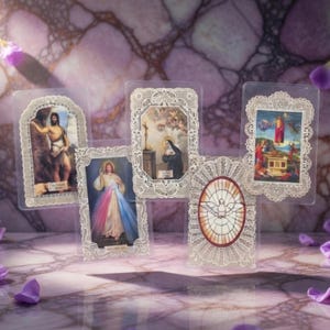 Puede incluir: Una colección de tarjetas de oración religiosas con varias figuras y escenas religiosas. Cada tarjeta está dentro de una funda de plástico transparente y enmarcada con delicado encaje blanco. Las imágenes representan a Jesús, santos y símbolos religiosos.