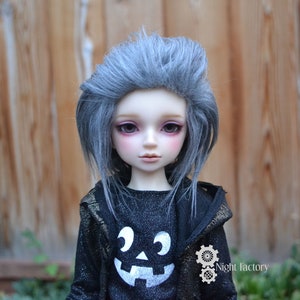 May include: A doll with gray hair and a black jacket with a pumpkin face design. The doll has a pale complexion, pink blush, and dark eye makeup. The jacket has a hood and a glittery texture. The doll is a Halloween-themed collectible.