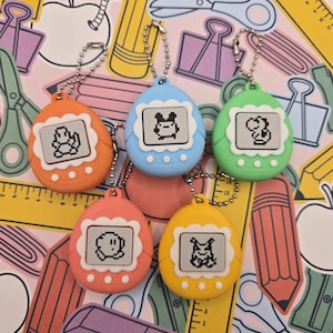 May include: A collection of colorful, retro-style keychain toys in orange, blue, green, coral, and yellow. Each keychain features a small screen displaying a pixelated character. The background is a pink pattern with school supplies.