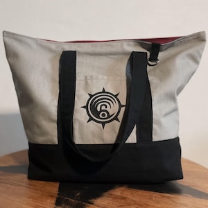 Op de afbeelding: Een canvas draagtas met een lichtgrijs bovenstuk en een zwarte onderkant. De tas heeft zwarte handvatten en een zwart grafisch ontwerp op de voorkant. De binnenvoering is rood. De tas staat op een houten tafel.