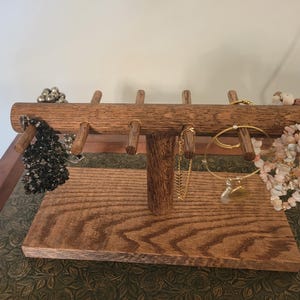 May include: A wooden jewelry display stand with a rectangular base and a horizontal bar with pegs. Several bracelets and necklaces in various colors and styles hang from the pegs. The stand is made of stained wood.