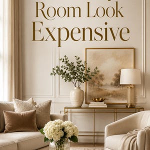 Op de afbeelding: Een woonkamer met een beige bank, bruine en textuurrijke kussens, een marmeren salontafel met een glazen vaas met witte hortensia's en een gouden kaars, en een gouden sidetable met een schilderij, lamp en vaas. Tekst: Make Any Room Look Expensive.