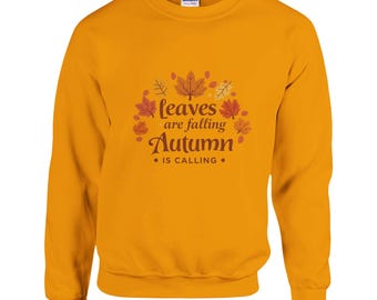 Puede incluir: Sudadera amarilla mostaza con el texto "leaves are falling Autumn is calling" en un diseño con temática otoñal. El diseño incluye hojas marrones y naranjas y elementos decorativos. La sudadera tiene cuello redondo y mangas largas.