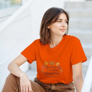 May include: An orange t-shirt with the text "Leaves are falling, Autumn is calling" in a fall-themed design. The shirt is being worn by a person sitting on steps. The shirt is a solid orange color.