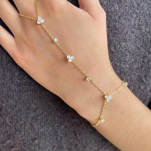 Könnte beinhalten: Ein zartes goldenes Handkettenarmband mit kleinen, klaren Edelsteinen und floralen Designs. Das Armband verbindet das Handgelenk mit dem Finger mit einer feinen Kette und schafft ein elegantes und feminines Accessoire.
