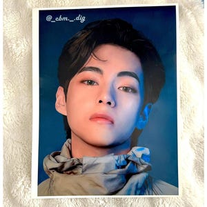 May include: A printed photograph of a person with dark hair and a neutral expression. The person is wearing a grey and blue jacket. The background is a blue gradient. The text "@_cbm._dig" is in the upper left corner.