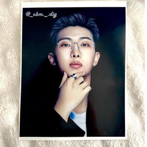 May include: A portrait print of a person wearing silver-rimmed glasses and a ring, dressed in a dark suit. The image includes the text "@_cbm._dig". The background is dark, and the print is on a white surface.