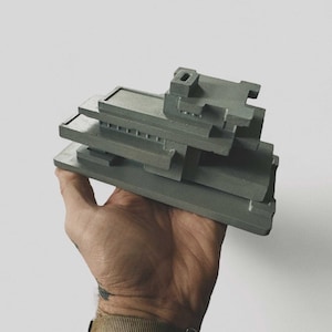 Könnte beinhalten: Graues Architekturmodell eines Gebäudes mit mehreren Ebenen und geometrischen Formen. Das Modell wird in einer Hand vor weißem Hintergrund gehalten. Das Gebäude hat ein flaches Dach und rechteckige Fenster.