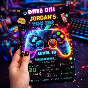 Peut inclure: Une carte d'invitation noire avec un design de manette de jeu aux couleurs néon. La carte indique "GAME ON! JORDAN'S, YOU IN?" et "LEVEL 10". Des graphiques supplémentaires incluent un hamburger, une canette de soda et des étoiles. La carte est pour une fête d'anniversaire.