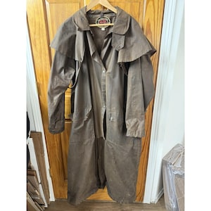May include: A long, brown, waxed canvas duster coat featuring a wide shoulder cape, a front button closure, and two large front pockets. The interior neck label displays the text The Australian Outback Collection.