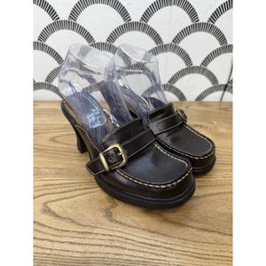 May include: A pair of dark brown leather mules featuring a square toe, a thick strap across the vamp with a brass-toned metal buckle, and contrast white stitching along the edges. The shoes have a mid-height heel and a black sole.