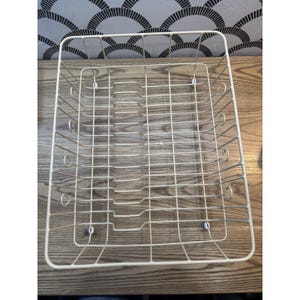 May include: Off-white rectangular wire dish rack. The rack has a grid pattern with curved edges and is designed to hold plates and other kitchen items. The rack is made of metal and sits on a wooden surface.