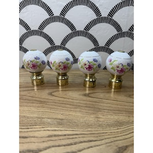 May include: Four spherical curtain rod finials featuring a white ceramic base with a floral pattern of pink, purple, and green flowers. Each finial is attached to a polished gold-tone metal base designed for mounting onto a curtain rod pole.