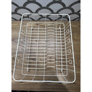 May include: A white, rectangular dish rack made of wire. The rack has a grid pattern with curved supports for plates and utensils. It sits on a wooden surface, with a patterned wall in the background.