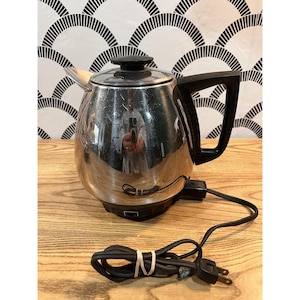 May include: A vintage, stainless steel electric coffee percolator with a black handle and lid. The coffee pot has a glass top and a black power cord. The percolator is sitting on a wooden surface.