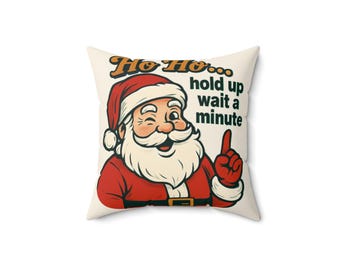 Puede incluir: Cojín cuadrado con un diseño festivo que presenta a Papá Noel guiñando un ojo y señalando. El texto dice "Ho Ho... hold up wait a minute". El cojín tiene un fondo blanco y una ilustración de Papá Noel en rojo y blanco.