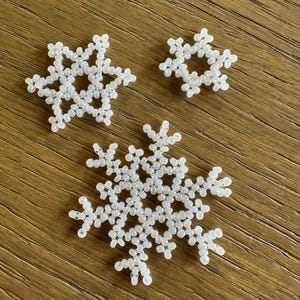 Pode incluir: Três enfeites de flocos de neve brancos com contas de tamanhos variados. Os flocos de neve são feitos de pequenas contas redondas e estão dispostos sobre uma superfície de madeira. O floco de neve maior está no centro, com dois menores acima.