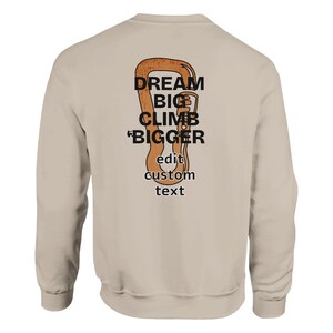 Könnte beinhalten: Beiges Sweatshirt mit großem orange-braunen Karabinerhaken-Motiv auf dem Rücken. Der Aufdruck enthält die Worte "DREAM BIG CLIMB BIGGER" und "edit custom text" in schwarzer Schrift.