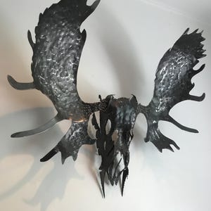 Puede incluir: Una gran escultura metálica de una cabeza de alce con cornamenta. La escultura está hecha de metal oscuro y texturizado y está montada en una pared blanca. La cornamenta es ancha y extendida, y la cabeza tiene una apariencia esquelética detallada.