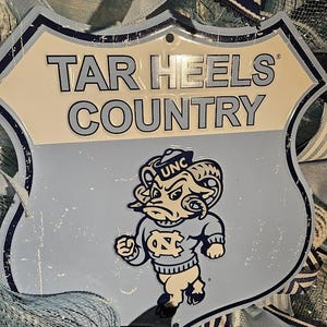 Pu&ograve; includere: Un cartello in metallo azzurro con le parole "TAR HEELS COUNTRY" in grassetto. Il cartello presenta un ariete dei cartoni animati con un maglione UNC. Il cartello ha la forma di uno scudo e ha un aspetto invecchiato.